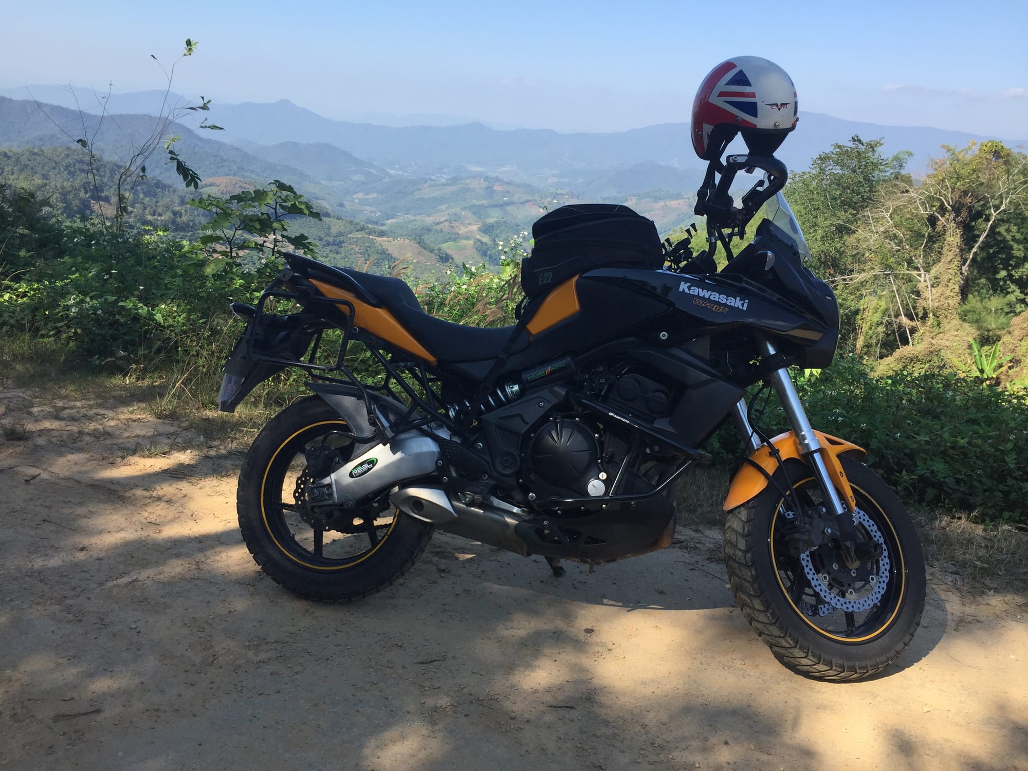 A Kawasaki Versys touring motorcycle parked in the mountainous Chiang Rai region of Northern Thailand, surrounded by lush tropical greenery.